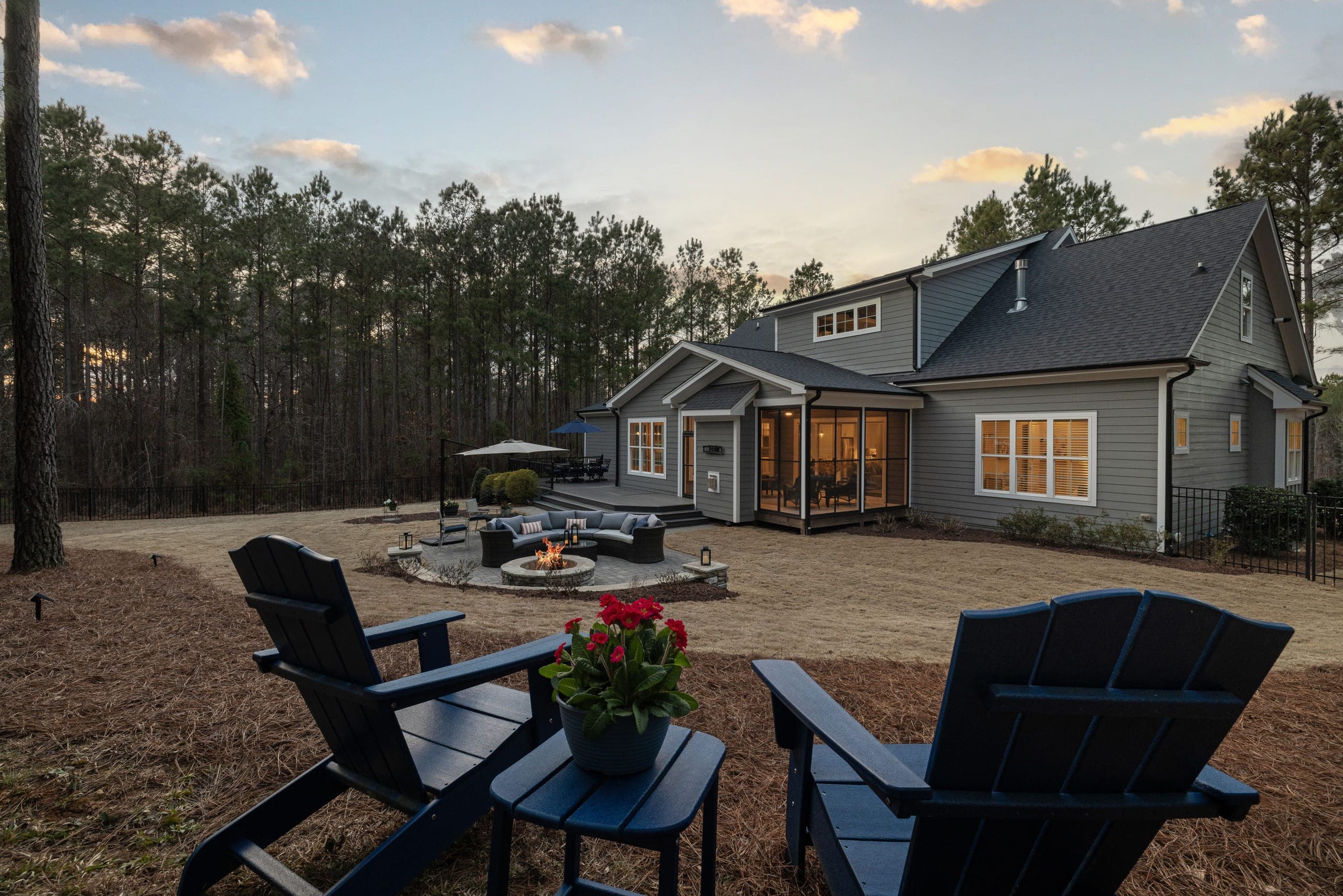 A backyard at dusk with a lit stone fire pit and curved seating on a paver patio, in front of a gray house with a glowing screened porch; two dark Adirondack chairs sit in the foreground.