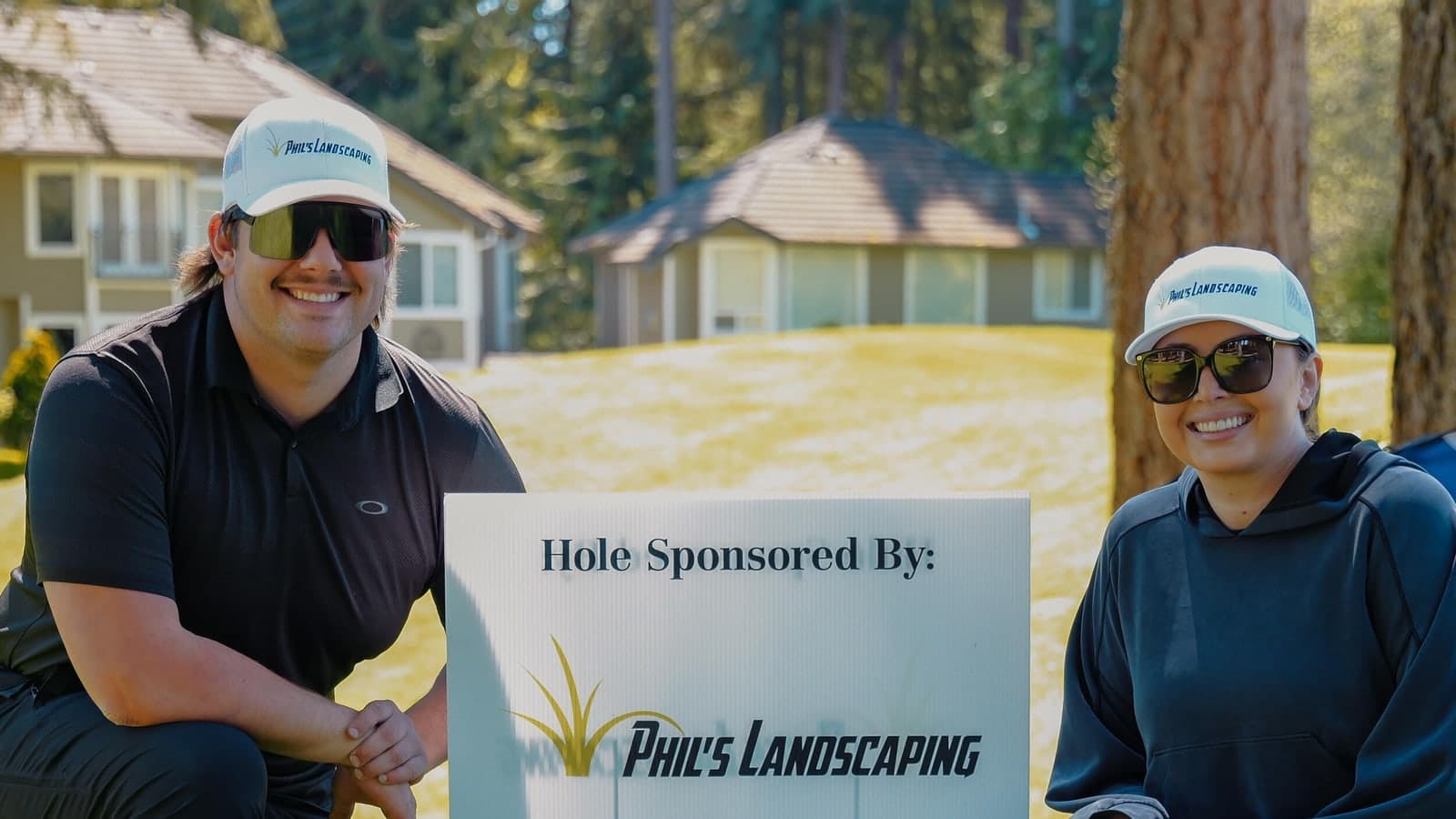 Two members of the Phil's Landscaping team kneeling beside a golf-tournament hole sponsor sign.