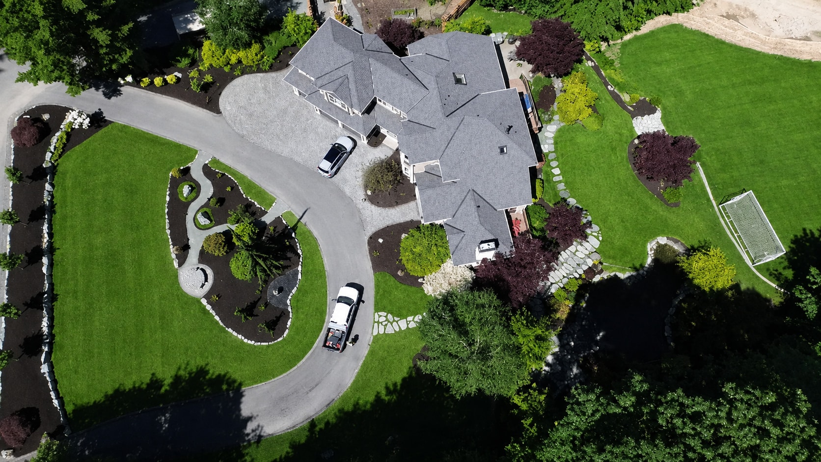 A drone view looking straight down on a large property: a curved driveway loops through striped lawn and stone-edged planting beds to a gray-roofed house.
