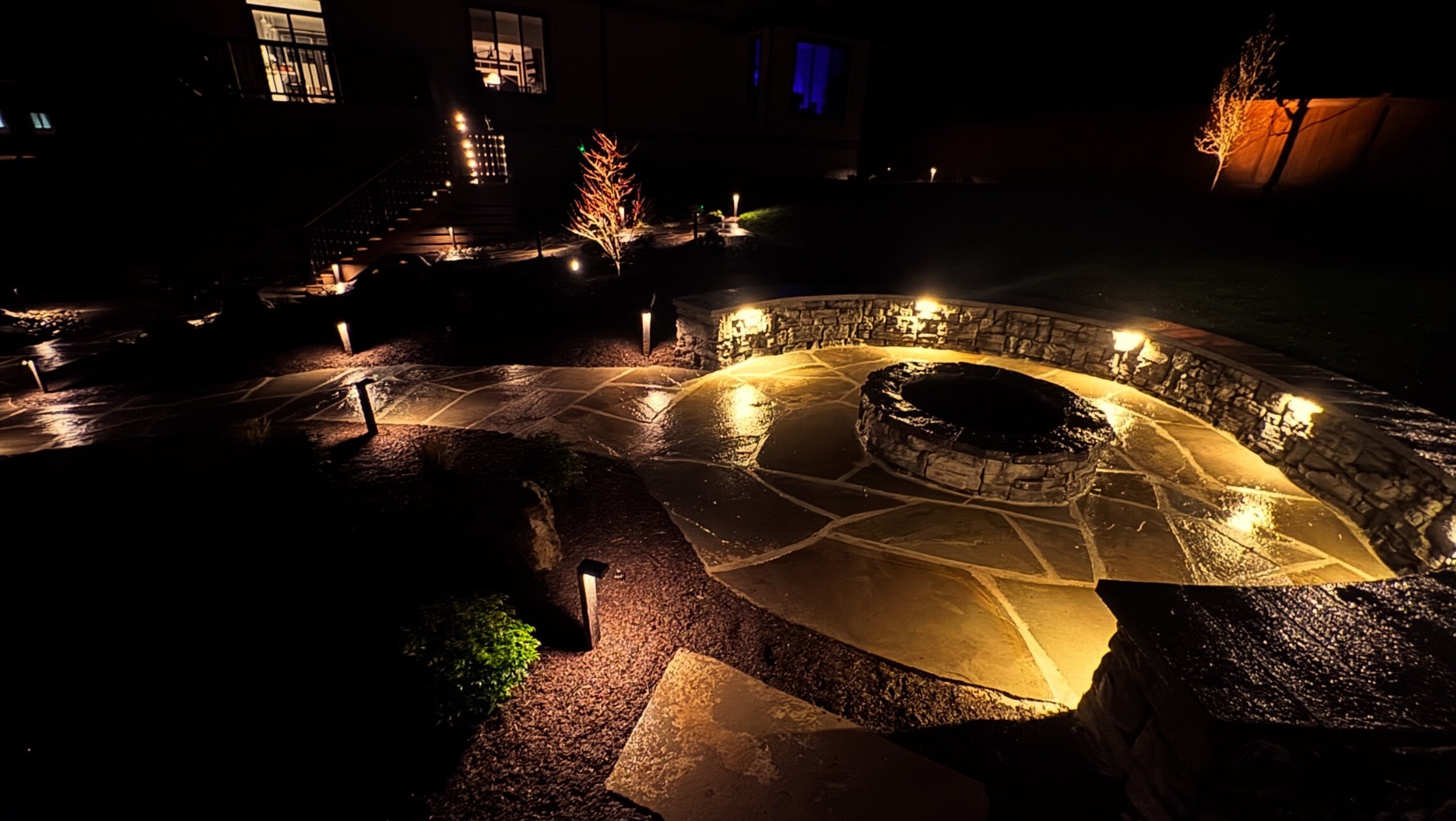 A flagstone patio at night around a round stone fire pit, ringed by a curved stone seat wall with built-in lights, with path lights and lit trees leading back to the house.