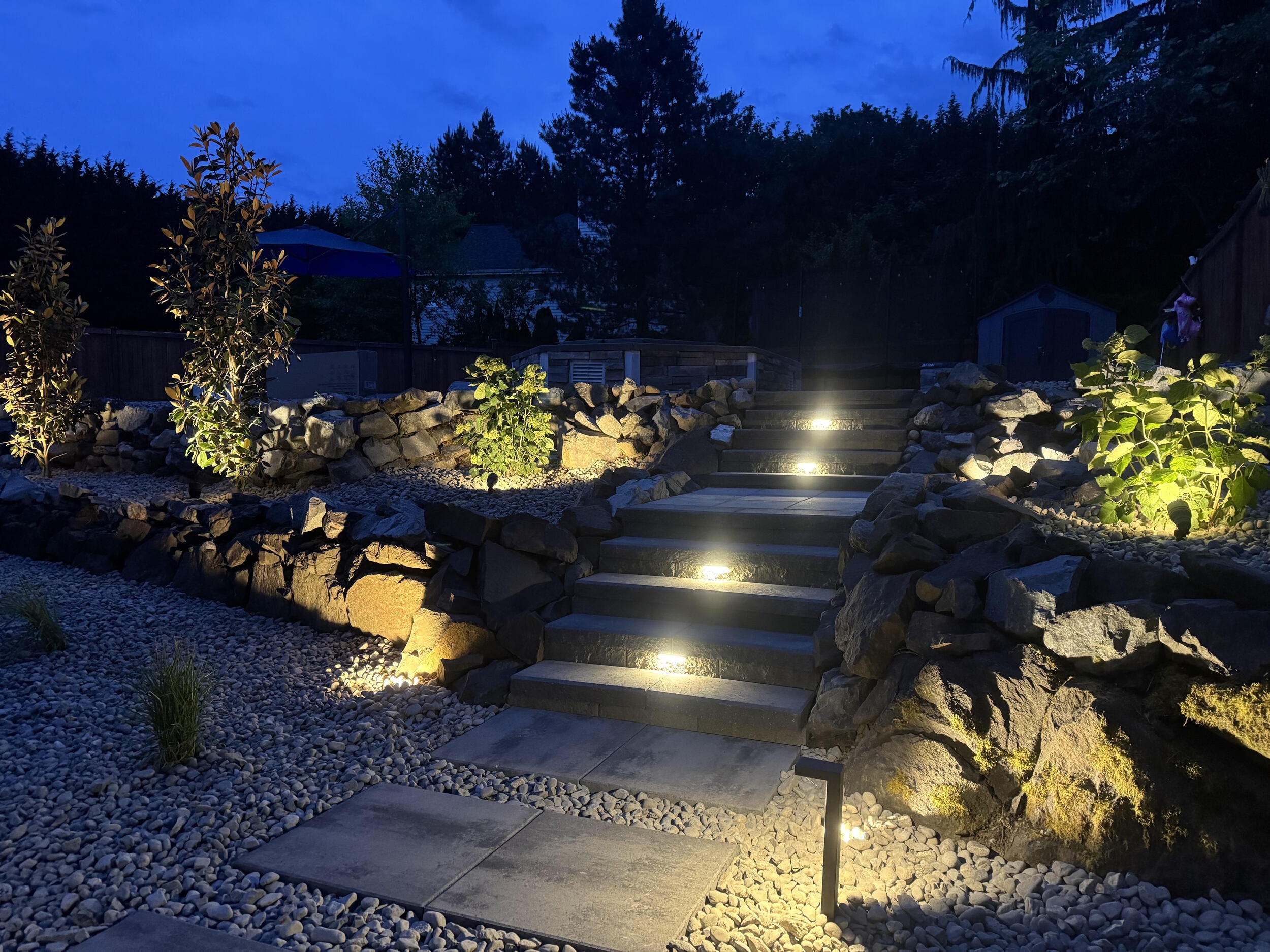 Wide stone steps with built-in step lights climbing between boulder walls and river-rock gravel at dusk, with up-lit shrubs on either side.