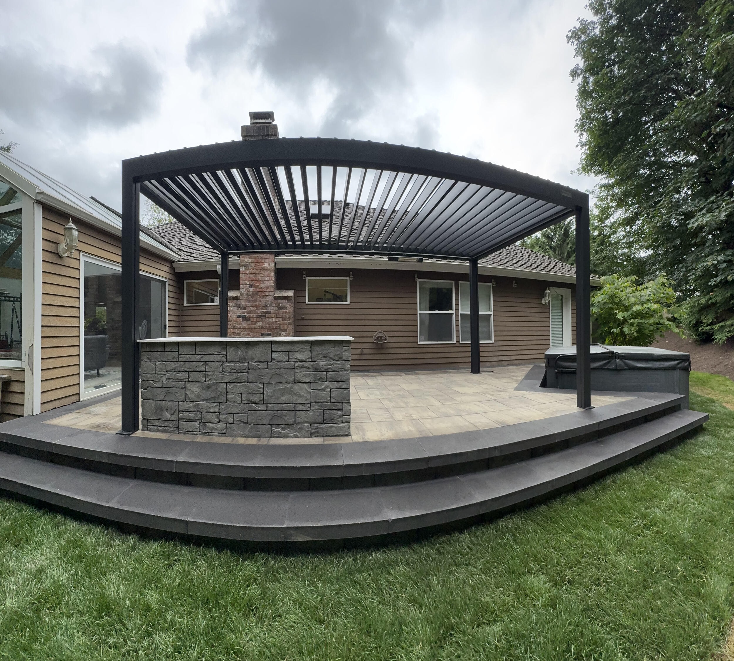 A louvered metal pergola over a raised paver patio with a stone-clad island counter, curved dark steps down to the lawn and a covered hot tub in the far corner, in front of a house with a brick chimney.
