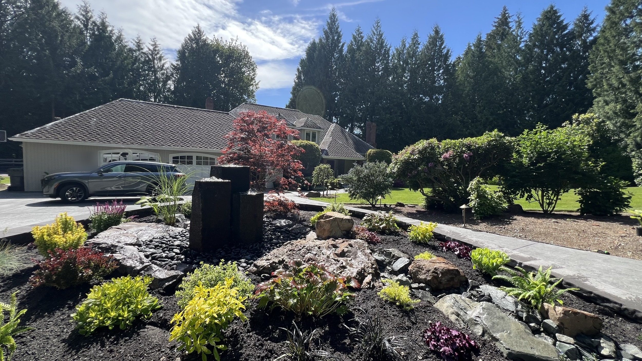 A sunny front garden bed with a water feature of three dark basalt columns over black river rock, a red Japanese maple, boulders and yellow-green and purple plantings, beside a paver walkway, with a gray house and a car in the driveway behind.