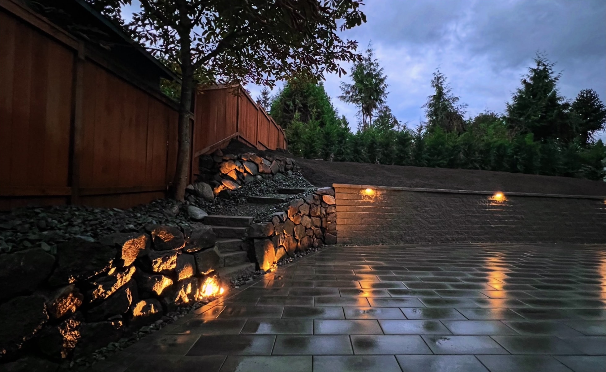 A boulder wall lit from below beside stone steps and a block retaining wall with cap lights, above a wet slab-paver patio at dusk, with a cedar fence and a tree on the left.